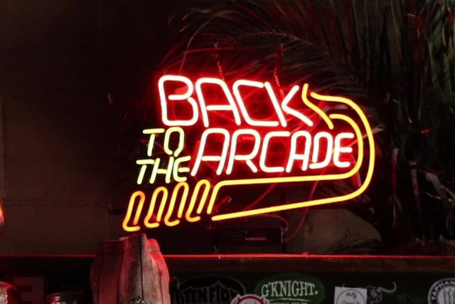 A red and yellow neon sign reading Back to the Arcade, mounted above the bar shelf