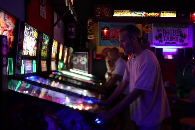 Two people playing pinball side by side in the dark backroom, playfields glowing under the machine lights