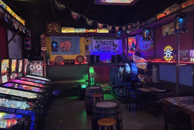 The backroom arcade — a row of lit pinball machines on the left, Pac-Man and Joust marquees overhead, Delirium and Rogue Farms banners on the wall, barrel tables in the middle