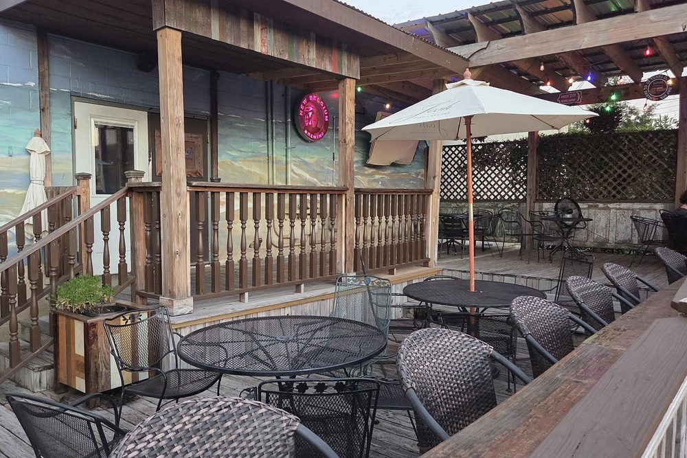 The Hang Loosa patio at dusk — wrought-iron tables and a white umbrella on the deck, string lights under the pergola, and a painted beach mural along the back wall