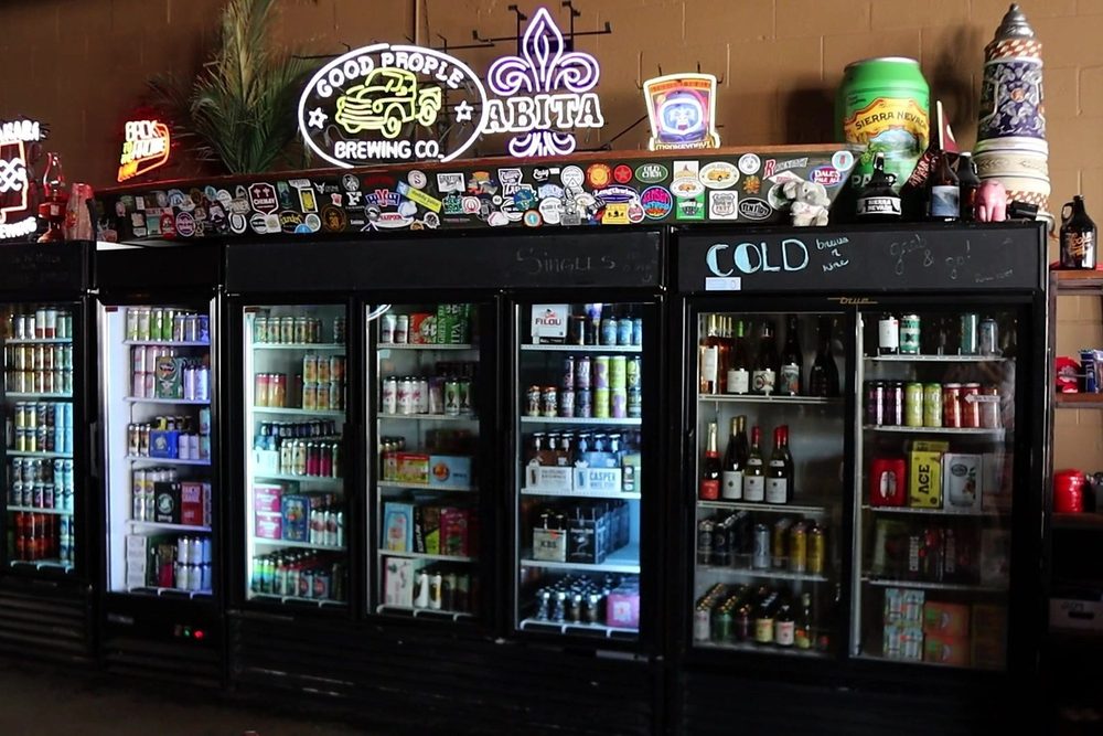 The to-go coolers at Loosa Brews — glass-door cases of cans, bottles and wine under neon Good People and Abita signs, with a sticker-covered rail above