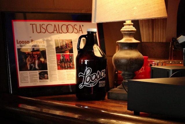 An amber Loosa Brews growler on the bar beside a lamp and a framed Tuscaloosa magazine feature about the shop