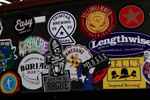 The sticker wall above the coolers, layered with brewery stickers — TrimTab, Yellowhammer, Rogue, Burial, Wiseacre, Lengthwise, Bell's